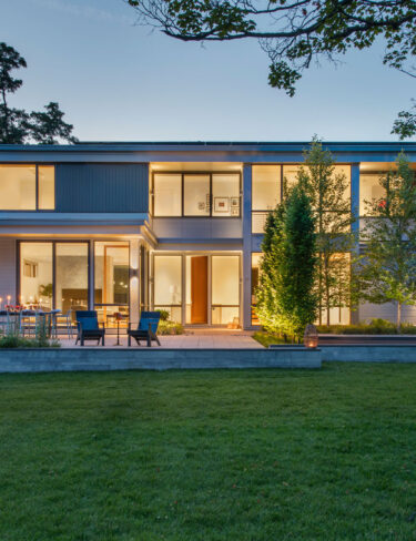Evening front house view warmly lit by interior and outdoor lighting alongside exterior walls.
