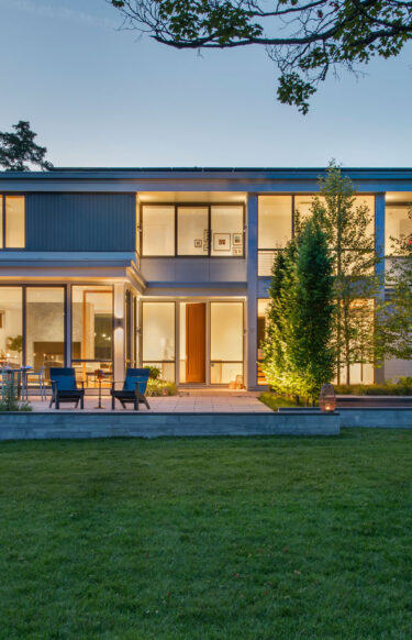 Evening front house view warmly lit by interior and outdoor lighting alongside exterior walls.